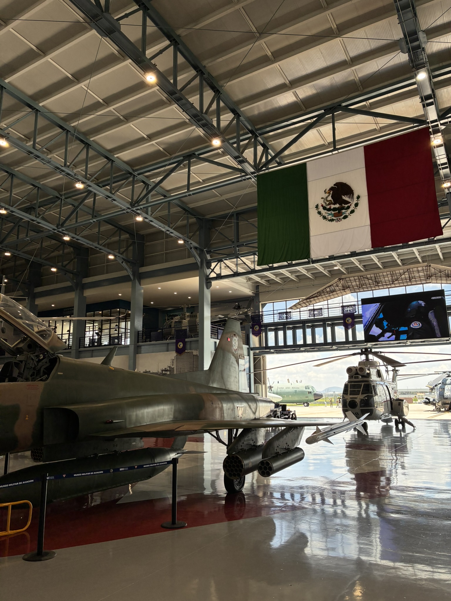 A military aviation museum hangar with a fighter jet and helicopter on display beneath a large Mexican flag