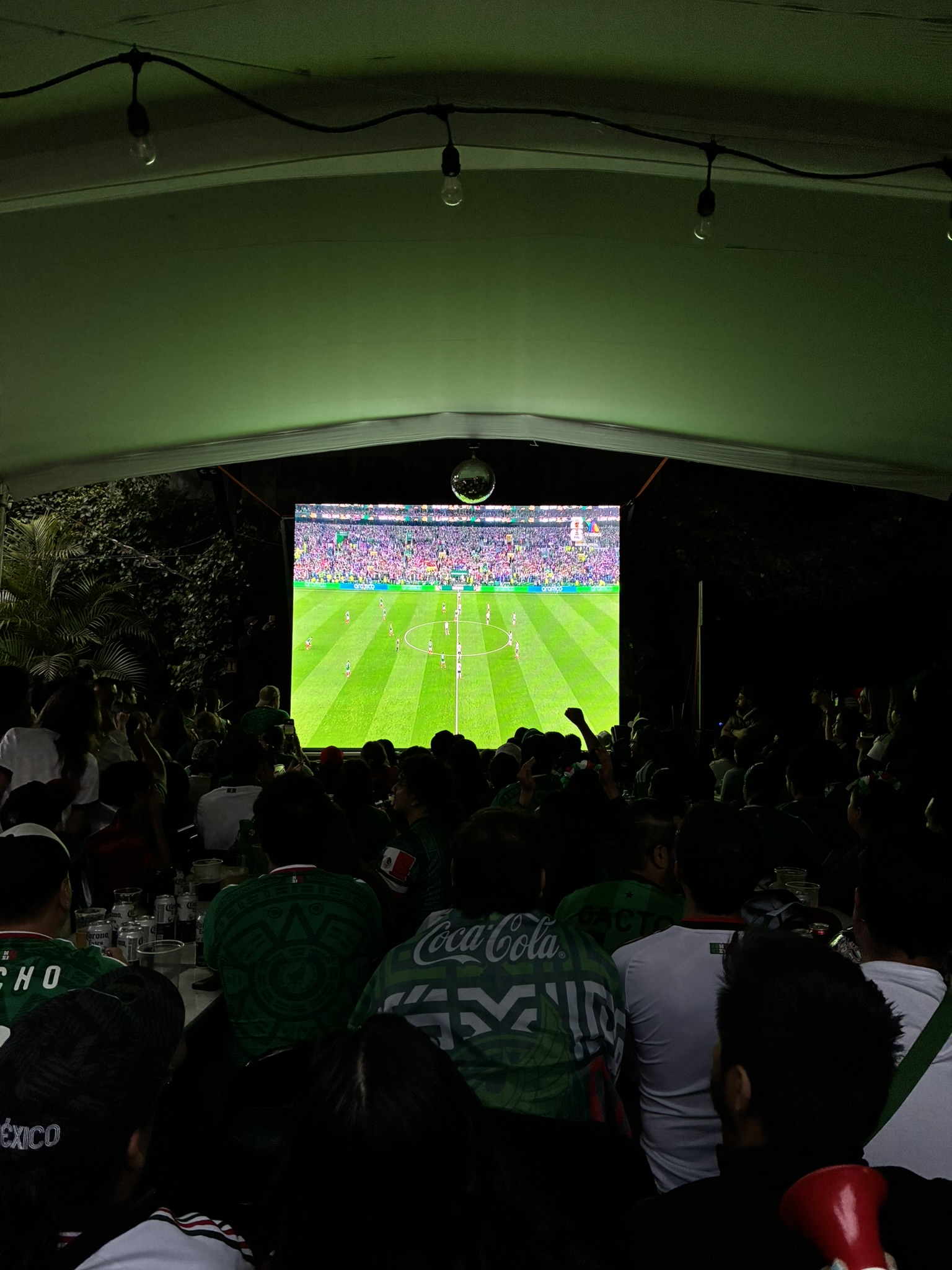 A crowd in green Mexico jerseys watching a match on a big screen at a bar