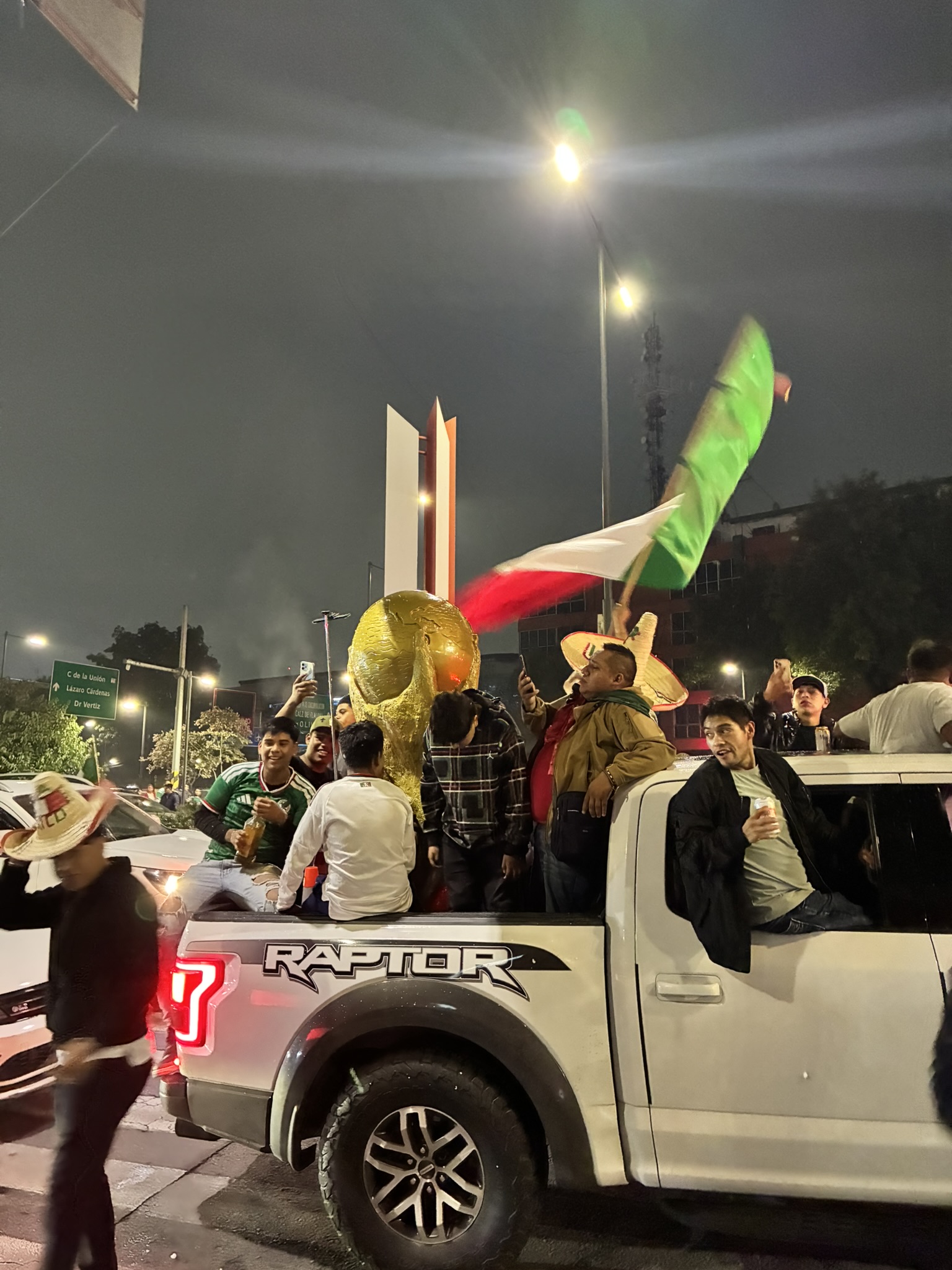People celebrating in the bed of a truck at night with a gold inflatable World Cup trophy and a waving Mexican flag