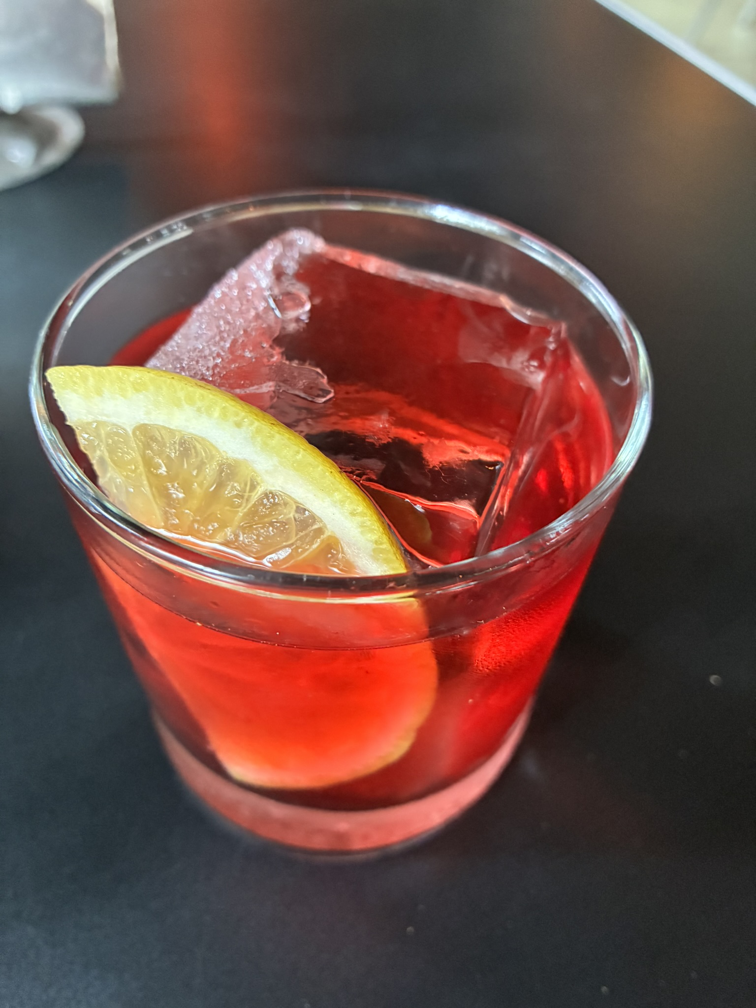 A red cocktail with a lemon wedge and large ice cube on a dark table