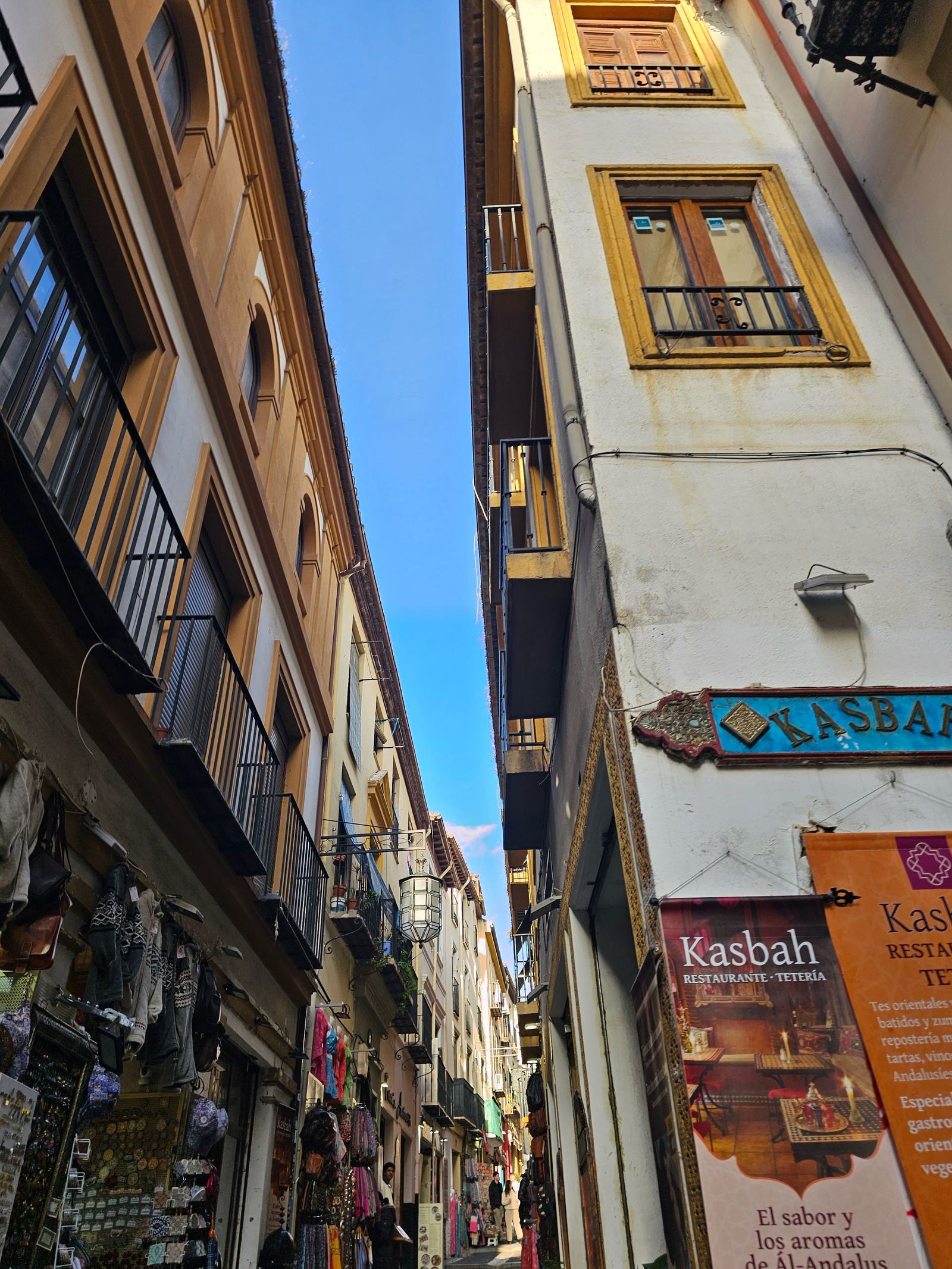 Looking up a narrow alley between tall buildings in Granada's Albaicín, with shop awnings and a strip of blue sky overhead