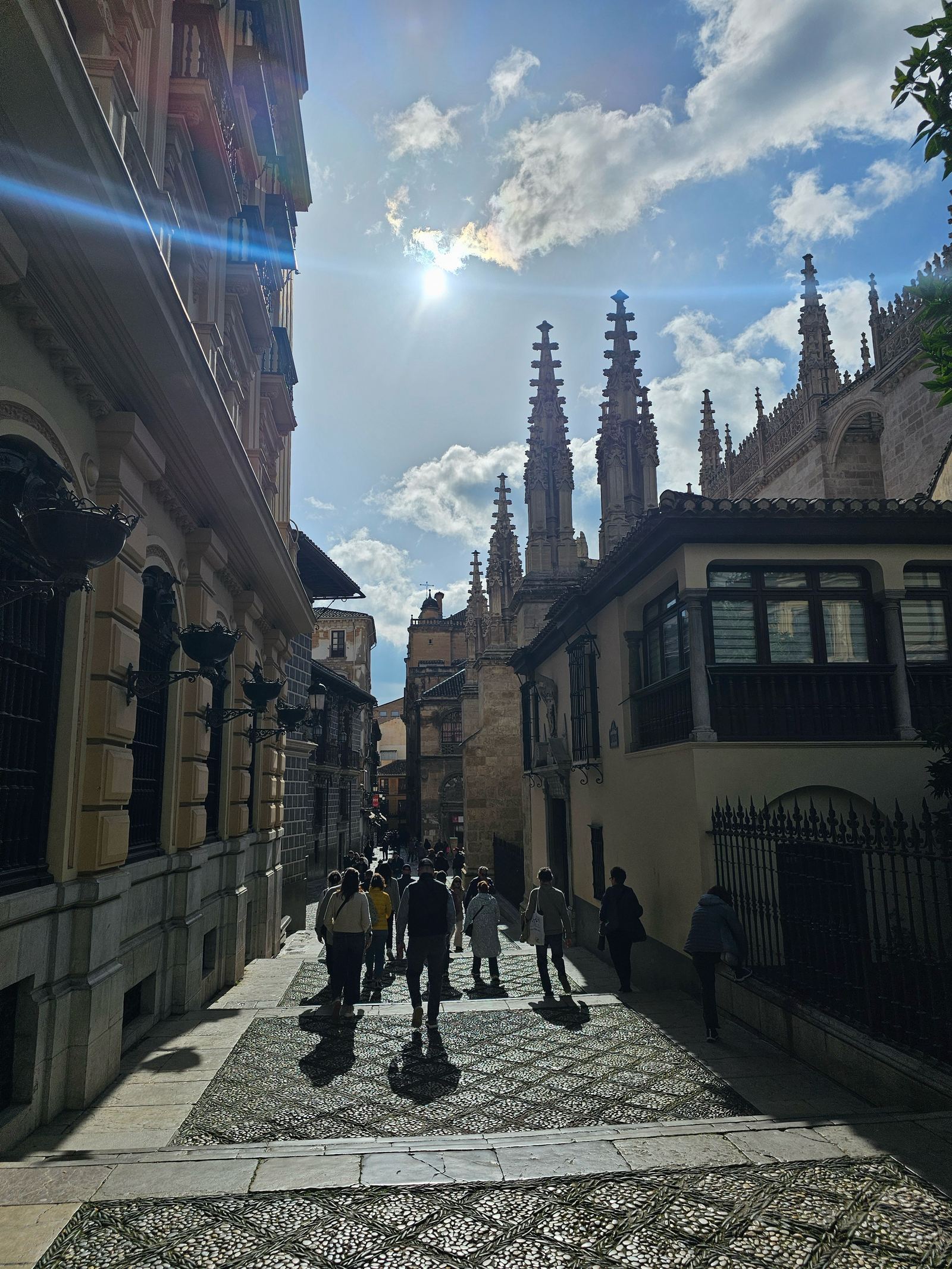 A pedestrian street in Granada leading toward the spires of the cathedral, with tourists walking beneath a sunny sky