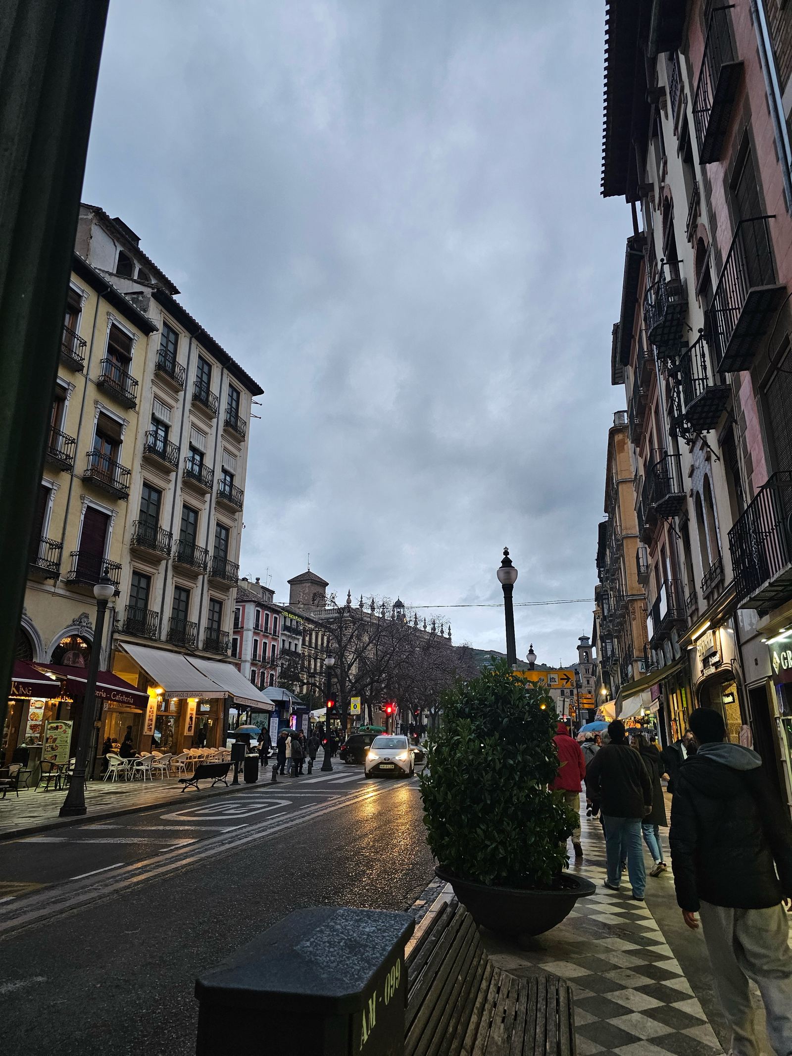 A rainy street in Granada at dusk with warm lights, a car, and pedestrians walking past cafés