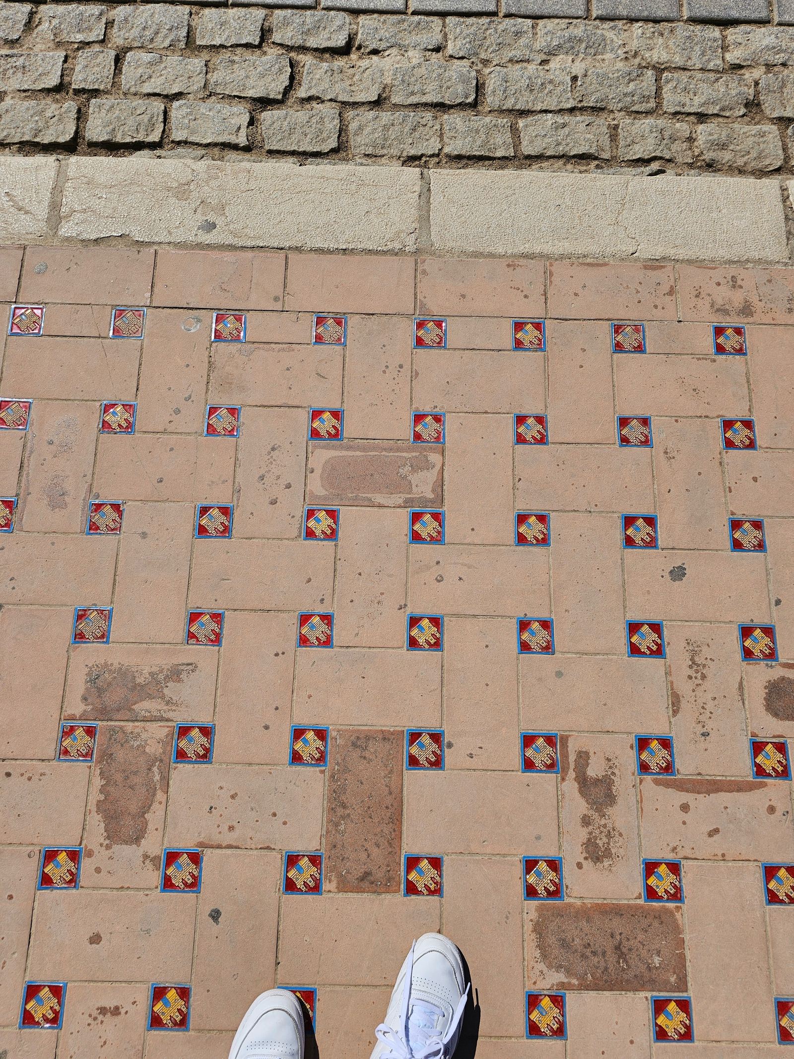 Looking down at a stone walkway in Sevilla decorated with small ceramic crest tiles, with sneakers visible at the bottom of the frame