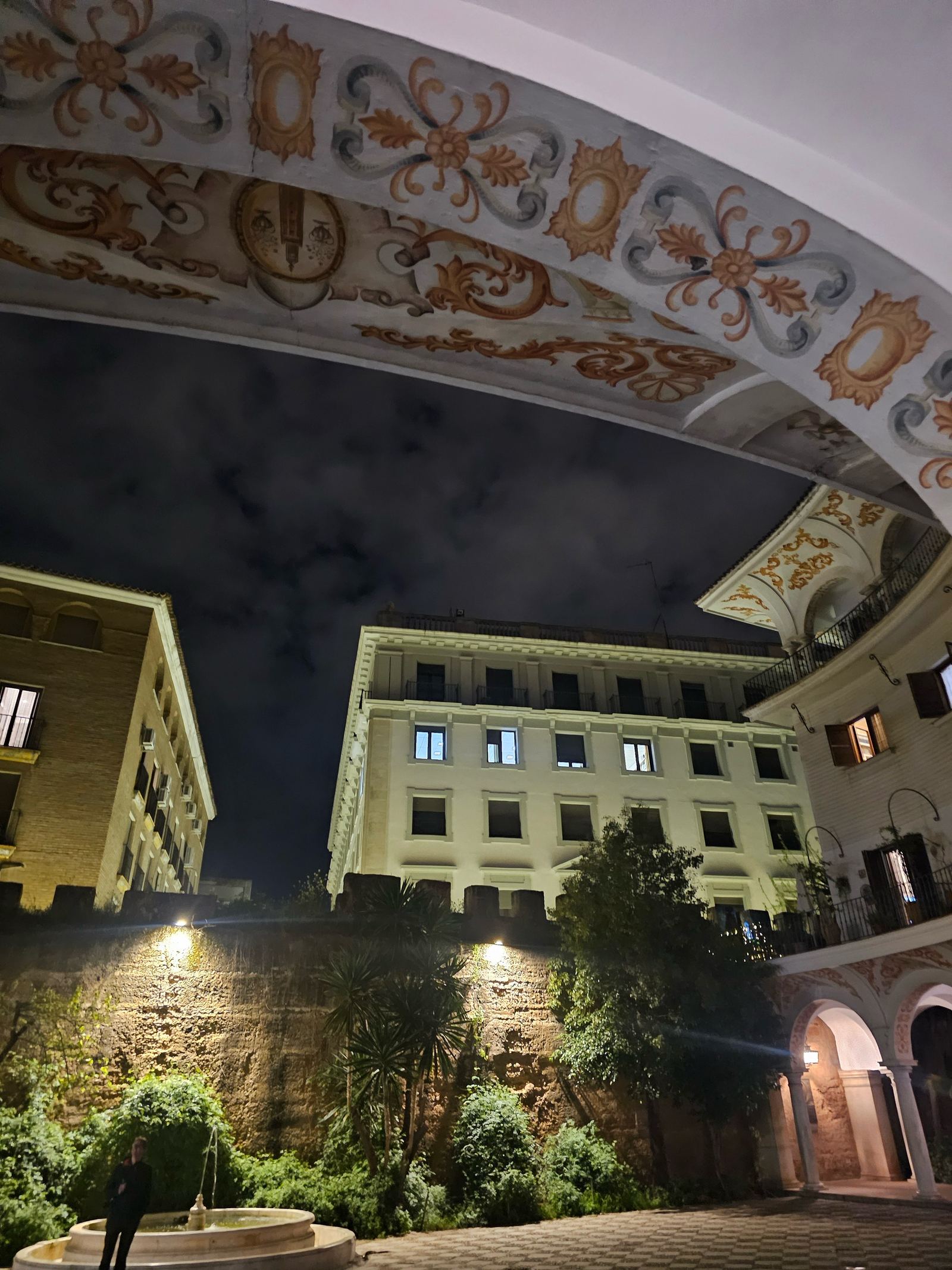 A courtyard in Sevilla at night, lit beneath an ornately painted archway with a fountain below